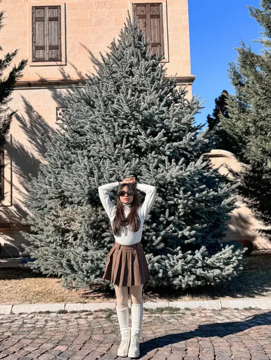 Fuji menenakan turtleneck putih, dipadu pleats mini skirt cokelat, legging beige, kaus kaki putih panjang, dan sepatu yang juga berwarna putih. Penampilannya semakin chic dengan sunglasses. [Foto: Instagram/fuji_an]