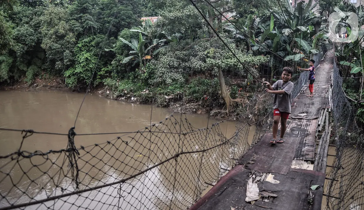 Anak-anak melintasi jembatan gantung yang sudah tidak layak di kawasan Srengseng Sawah, Jakarta, Sabtu (12/6/2021). Jembatan gantung tersebut hanya mengandalkan ikatan tambang besi yang mengait pada beton dan pepohonan yang ada di sekitar kali. (merdeka.com/Iqbal S. Nugroho)