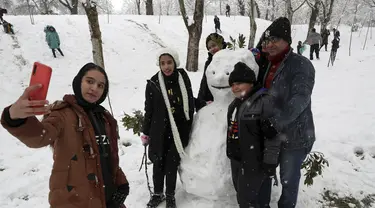 Orang-orang mengambil berswafoto dengan manusia salju ketika mereka menikmati salju di sebuah taman di Teheran utara, Iran (19/1/2020). Salju tebal menutupi jalan-jalan Teheran, menyebabkan penundaan penerbangan dan munutup sekolah, kata pihak berwenang di ibukota Iran. (AP Photo / Vahid Salemi)