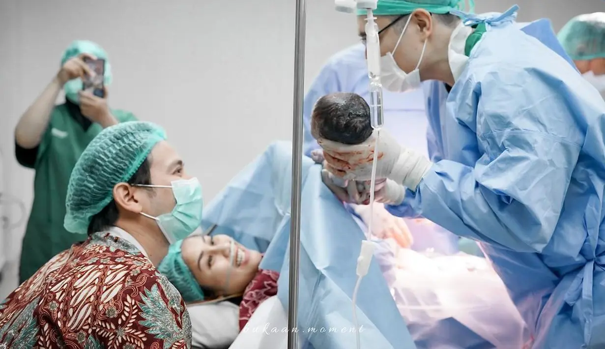 Pada foto lain, seorang dokter memegangi bayi. Dokter berbaju dinas lengkap plus sarung tangan dan penutup kepala memperlihatkan bayi yang baru lahir kepada Gya Sadiqah dan Tarra Budiman. Gya yang mengenakan alat bantu napas tersenyum semringah melibat putri keduanya. Sementara Tarra Budiman yang pakai masker tampak bahagia. Matanya berbinar melihat si kecil. (Foto: Bukaan Moment dari Instagram @gyaps)