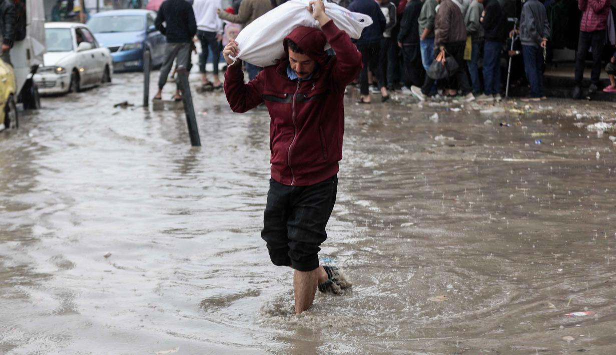 Kondisi tersebut semakin memperburuk krisis kemanusiaan yang dihadapi ratusan ribu pengungsi di Gaza. Tampak dalam foto, seorang pria membawa karung sambil berjalan di tengah banjir di kamp darurat yang menampung warga Palestina yang terlantar setelah hujan deras di Kota Gaza pada Selasa 25 November 2025. (Omar AL-QATTAA/AFP)