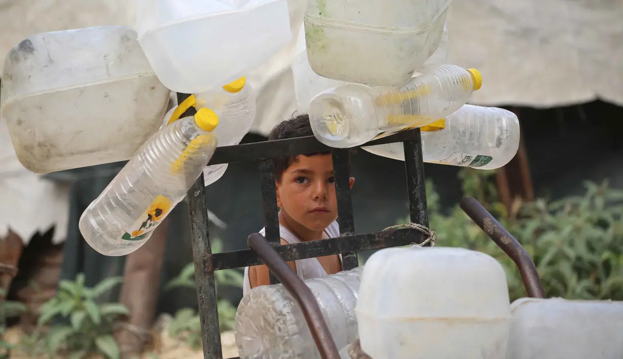 Seorang anak laki-laki Palestina berdiri di samping wadah plastik di atas troli, saat ia dan yang lainnya mengumpulkan air minum untuk keluarganya yang mengungsi dan tinggal di tempat penampungan setelah rumah-rumah mereka hancur akibat bombardir Israel, di kamp pengungsi Bureij, Jalur Gaza Tengah pada Senin 18 Mei 2026. (Eyad Baba/AFP)