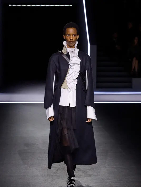 Kemeja transparan yang ringan kontras dengan mantel hitam tebal yang membentuk siluet tubuh. Jaket maskulin dipadukan dengan bustier, sementara coat tail dan kerah renda menjadi pelengkap yang menonjol.   [Dok/Dior],