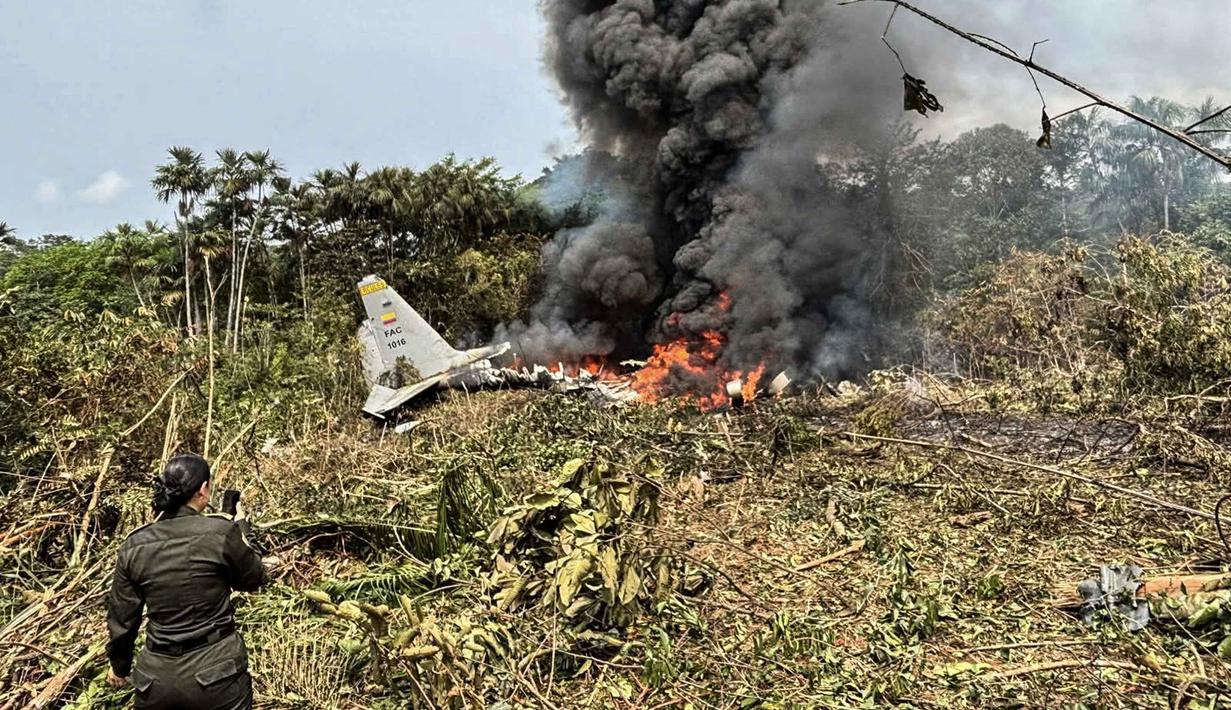 Pesawat berjenis C-130 Hercules itu jatuh tak lama setelah lepas landas dari Puerto Leguizamo, Kolombia, menyebarkan puing-puing yang terbakar di dasar hutan. Tampak dalam foto, kobaran api dan asap hitam tebal membubung dari pesawat Hercules Angkatan Udara yang jatuh saat lepas landas, sementara seorang anggota Kepolisian Kolombia berdiri di dekatnya, di Puerto Leguizamo, Kolombia, pada Senin 23 Maret 2026. (Daniel Ortiz/AFP)
