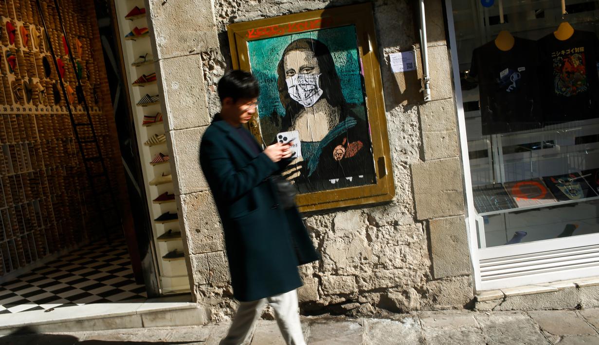 Seorang pria melewati poster seniman Italia TVBOY bergambar Mona Lisa karya Leonardo da Vinci mengenakan masker dan memegang smartphone di Barcelona, Selasa (18/2/2020). Instalasi muncul setelah Mobile World Congress (MWC) 2020 batal digelar karena wabah virus corona yang mencekam. (PAU BARRENA/AFP)