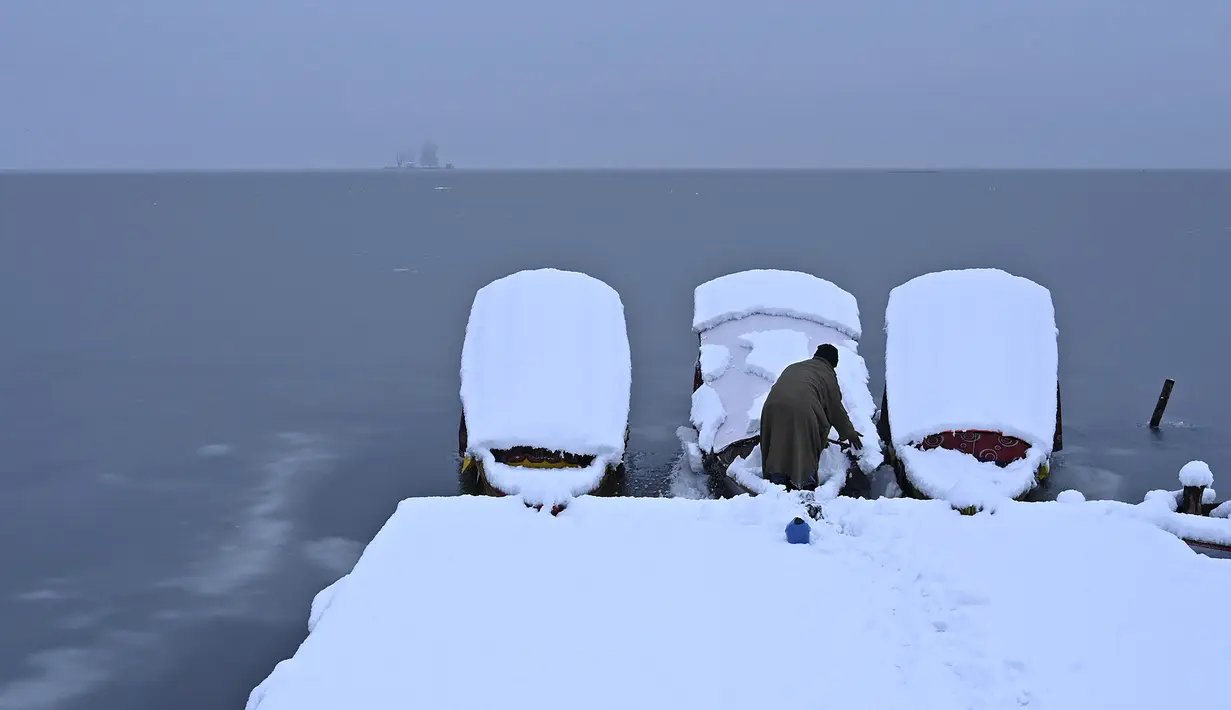 Seorang lelaki menyingkirkan salju dari sebuah kapal dekat bagian beku danau Dal setelah hujan salju lebat di Srinagar (16/1/2020). Temperatur telah turun beberapa tingkat di bawah titik beku. (AFP Photo/Tauseef Mustafa)