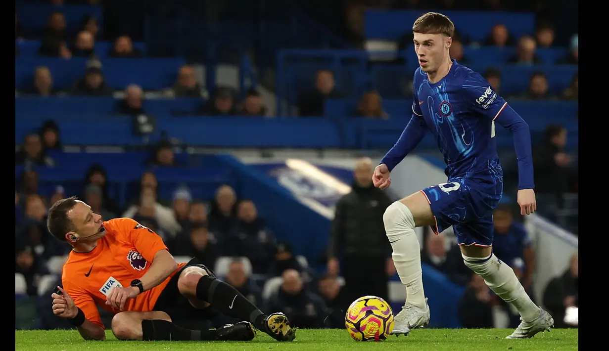 Pemain Chelsea, Cole Palmer (kanan) menggiring bola melewati wasit Stuart Attwell yang sedang terjatuh dalam laga lanjutan Liga Inggris 2024/2025 melawan West Ham di Stamford Bridge, London, Inggris, Selasa (04/02/2025) WIB. (AFP/Adrian Dennis)