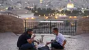 Dua pria Pelestina berbuka puasa selama bulan suci Ramadan dengan latar belakang Kota Tua Yerusalem dan kompleks Masjid al-Aqsa yang ditutup selama krisis pandemi coronavirus yang baru di Bukit Zaitun (19/5/2020). (AFP/Ahmad Gharabli)