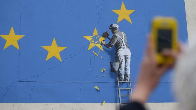 Warga mengambil gambar mural seorang pria yang tengah menghancurkan salah satu dari 12 bintang kuning bendera Uni Eropa di dinding kawasan Dover, Inggris, Senin (8/5). Mural karya seniman jalanan Banksy itu berjudul 'Brexit'. (DANIEL LEAL-OLIVAS/AFP)