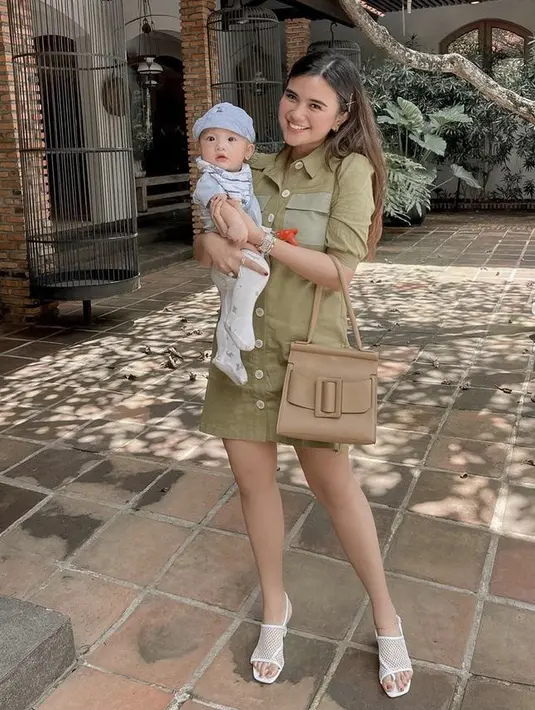 Sambil menggendong si kecil, Audi mengenakan mini dress hijau army dengan detail kancing depan dan  dua kantung. Ia pun membawa handbag cokelat dan heels putihnya.@audimarissa