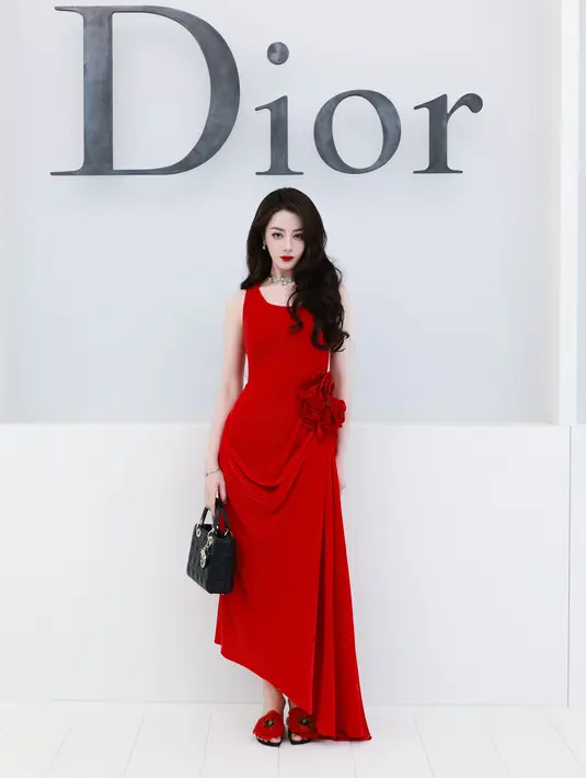 Dilraba mencuri perhatian dengan gaun beludru merah asimetris. Dihiasi bros bunga besar di pinggang, dipadukan tas Lady Dior hitam dan heels bermotif bunga merah [dok. Dior]