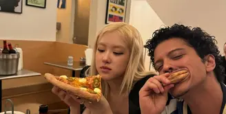 Sementara itu, Bruno Mars tampil dengan gaya playful khasnya. Ia mengenakan jaket varsity biru dengan aksen putih, dipadukan dengan kalung mutiara yang menjadi ciri khas penampilannya yang penuh karakter. Bruno tampak santai dan ceria, menggigit bagian crust pizza sambil tersenyum, memperlihatkan suasana akrab di antara mereka. [@roses_are_rosie].