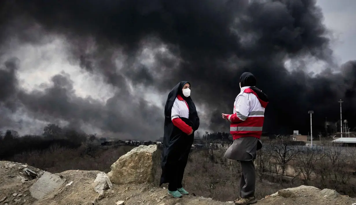 Beberapa laporan menyebut, adanya kebakaran besar di fasilitas kilang di ibu kota Iran, Teheran, setelah dihantam serangan udara pada Sabtu 7 Maret 2026 malam. Tampak dalam foto, dua wanita dari Palang Merah Iran berdiri saat kepulan asap tebal dari serangan AS-Israel terhadap fasilitas penyimpanan minyak Sabtu 7 Maret 2026 malam membubung ke langit di Teheran, Minggu, 8 Maret 2026. (AP Photo/Vahid Salemi)