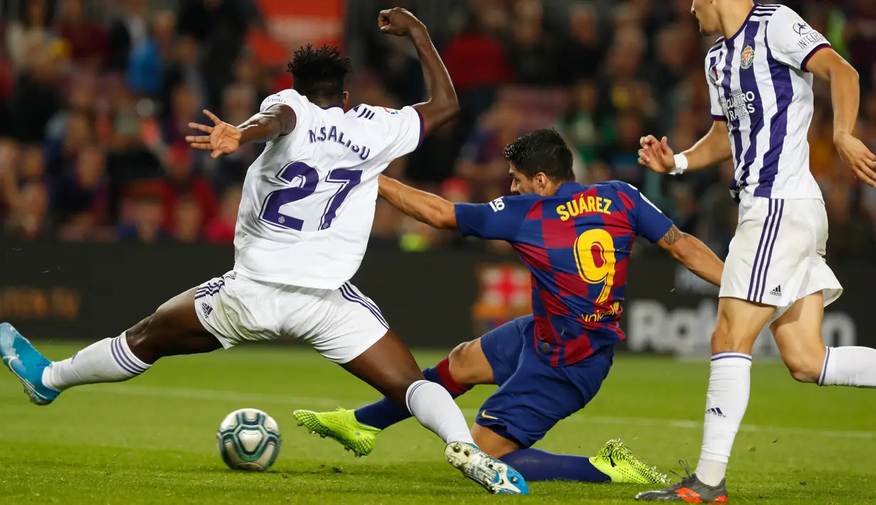 Striker Barcelona, Luis Suarez, menendang bola dibayangi pemain Real Valladolid, Mohammed Salisu dalam pertandingan pekan ke-11 La Liga  di Camp Nou, Selasa (29/10/2019). Barcelonamenutup laga dengan kemenangan 5-1 lewat gol tambahan dari Lionel Messi dan Luis Suarez. (AP/Joan Monfort)