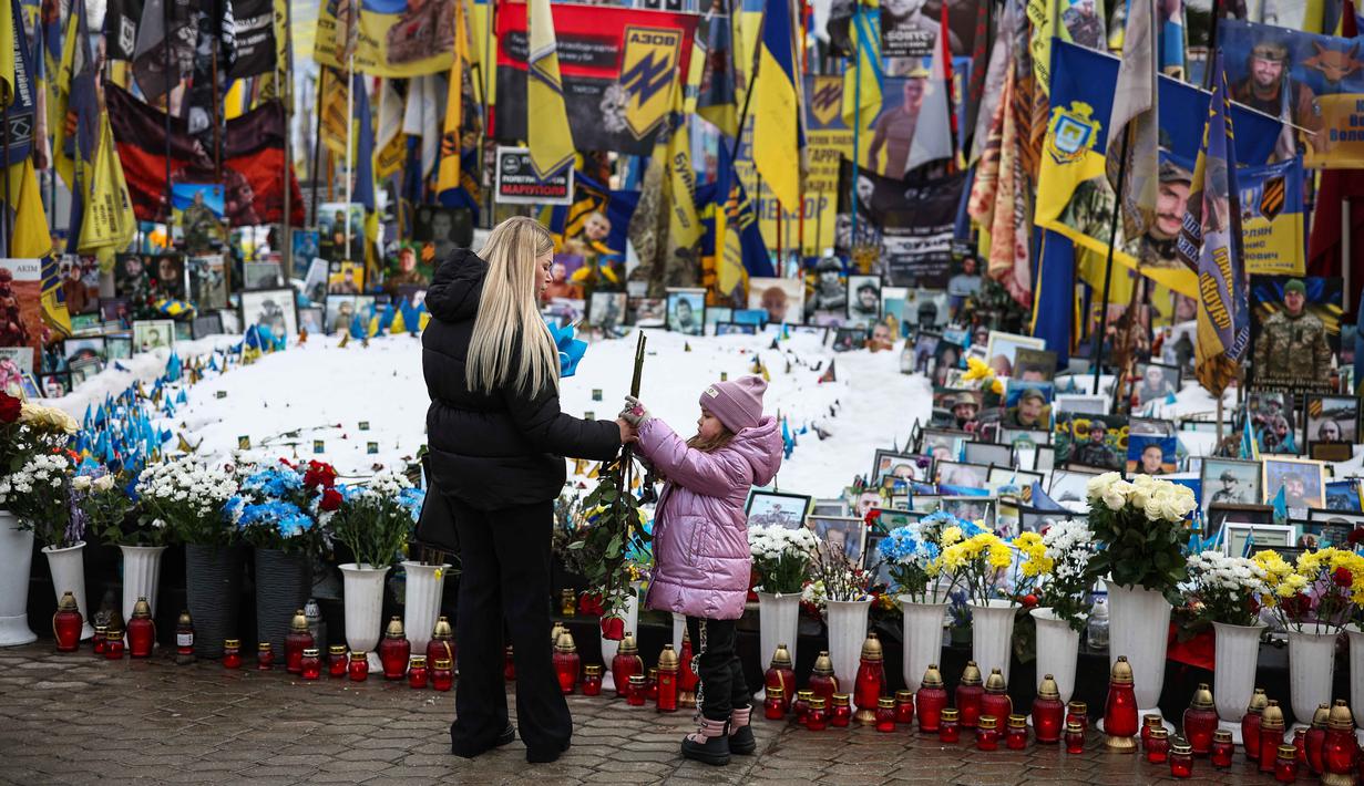 Ribuan tentara dari kedua pihak tewas di medan perang, sementara warga sipil Ukraina bertahun-tahun hidup di bawah ancaman serangan udara, pemadaman listrik, dan krisis air bersih. Tampak dalam foto, seorang wanita dan seorang anak mengunjungi tugu peringatan sementara untuk tentara Ukraina dan asing di Lapangan Kemerdekaan, Kyiv, saat peringatan keempat invasi Rusia ke Ukraina, Selasa 24 Februari 2026. (HENRY NICHOLLS/AFP)