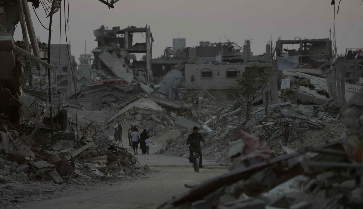 Penghalangan akses dan birokrasi ini jelas menghambat proses pemulihan dan memperpanjang penderitaan warga sipil Gaza. Tampak dalam foto, pengungsi Palestina berjalan di antara reruntuhan bangunan yang hancur di Khan Younis, Jalur Gaza, Sabtu 8 November 2025. (AP Photo/Abdel Kareem Hana)