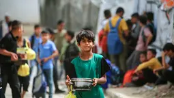 Seorang anak laki-laki Palestina yang mengungsi membawa sepanci makanan yang dikumpulkan dari dapur amal ke tempat penampungan keluarganya, di kamp pengungsi Bureij, Jalur Gaza Tengah pada Senin 18 Mei 2026. (Eyad Baba/AFP)
