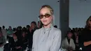 Emilia Clarke tampil dengan atasan zip abu-abu muda, dipadukan rok abu-abu gelap. Kacamata cokelat, tas Prada putih, dan heels slingback hitam lengkapi busananya [dok. Prada]