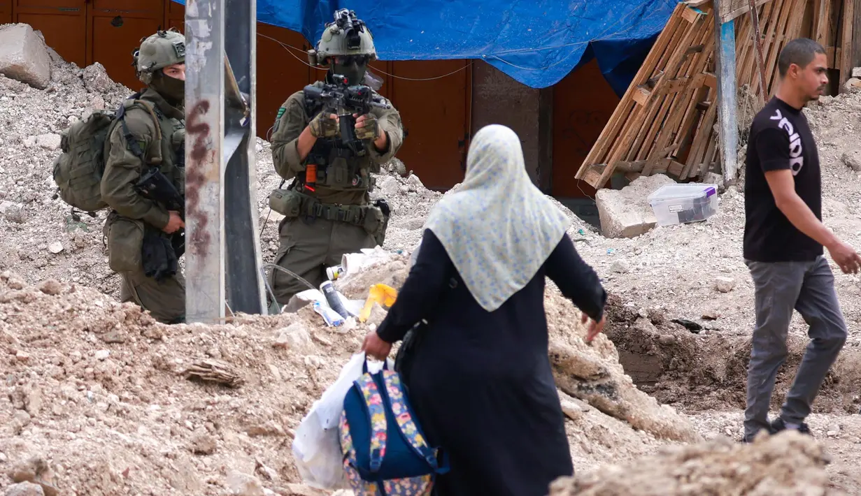 Seorang tentara Israel mengangkat senapannya saat warga Palestina membawa barang-barang mereka setelah diperintahkan untuk meninggalkan rumah mereka selama operasi Israel di kamp pengungsi Tulkarem di barat laut Tepi Barat yang diduduki pada 2 Mei 2025. (Jaafar ASHTIYEH/AFP)