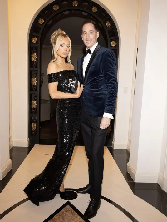 Setelah menjadi orangtua, Paris tetap tampil glamor mengenakan gaun off shoulder sqeuin hitam. Dan sang suami dengan jas velvet birunya.(@parishilton)