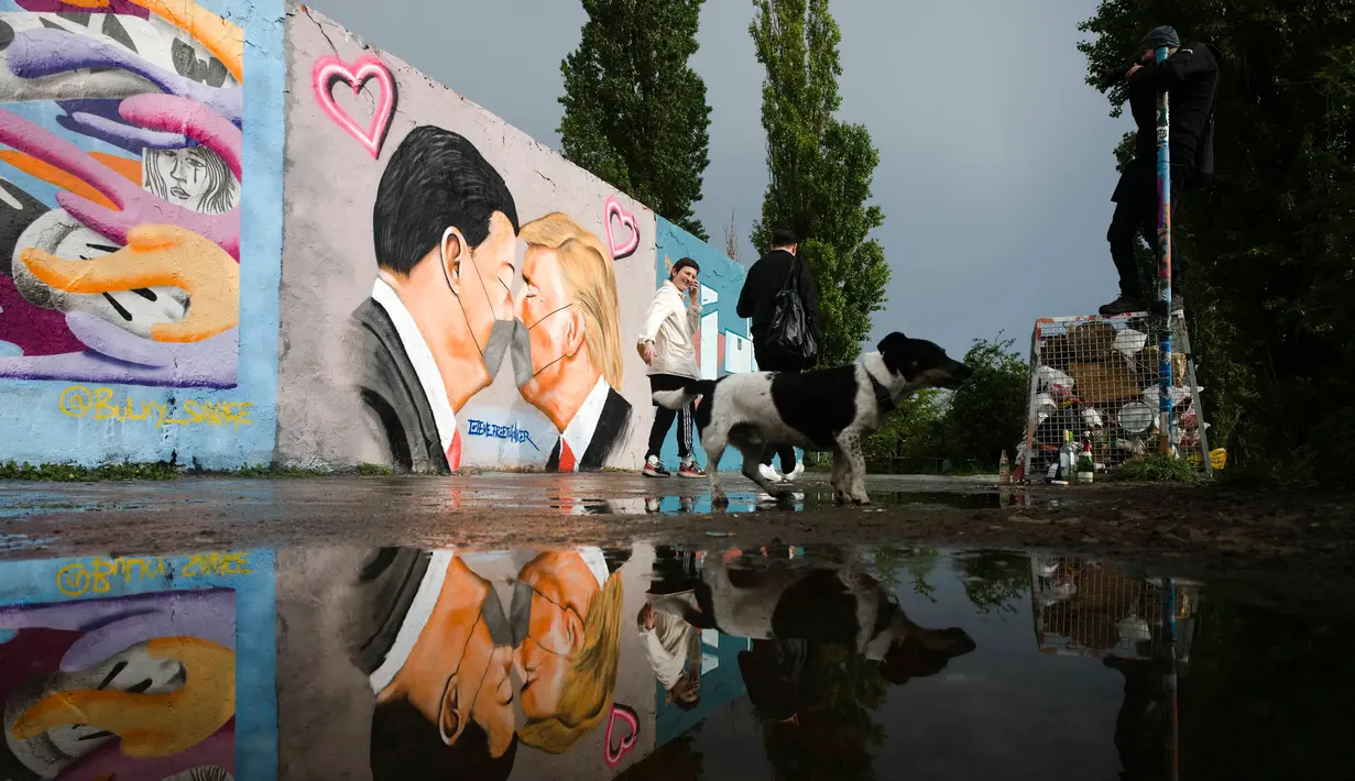 Orang-orang berjalan di depan Graffiti yang menggambarkan Presiden AS Trump (kanan) dan Presiden Tiongkok Xi Jinping saling berciuman dengan mengenakan masker di tembok taman umum Mauerpark di Berlin, Jerman, Rabu, (29/4/2020). (AP/Markus Schreiber)