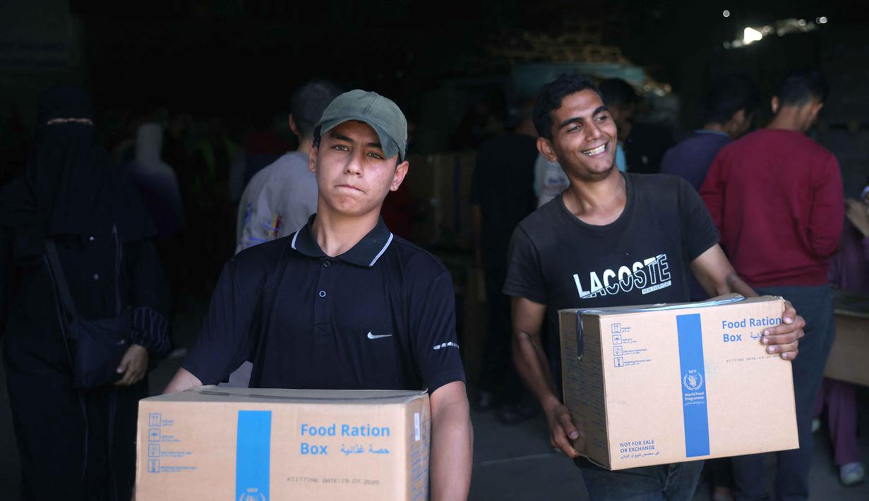 Dua pria Palestina masing-masing membawa kotak makanan yang disediakan oleh Program Pangan Dunia (WFP), yang diambil di pusat distribusi di desa Al-Zawayda, di tengah-tengah Jalur Gaza pada 27 Oktober 2025. (BASHAR TALEB/AFP)