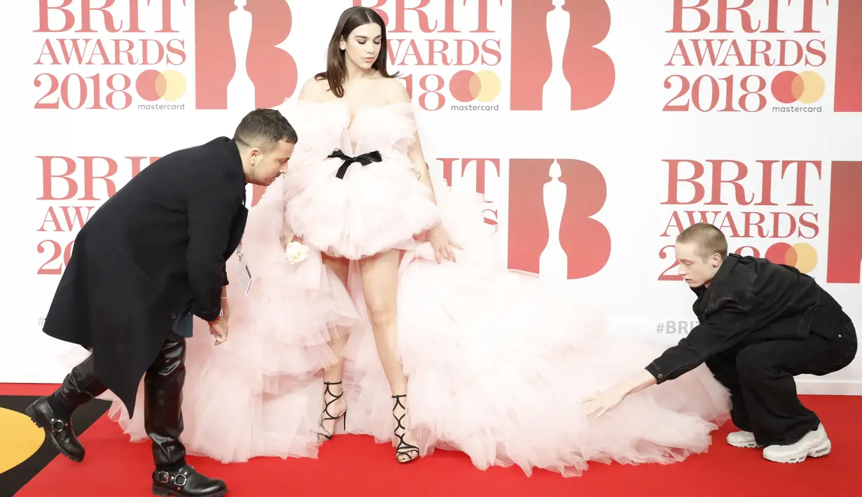 Dua orang pria membantu penyanyi cantik Dua Lipa merapikan gaunnya sebelum berpose di karpet merah acara Brit Awards 2018 di O2 Arena, London, Rabu (21/2). Ini adalah gelaran ke-38 untuk ajang penghargaan terbesar di Britania Raya. (Tolga AKMEN / AFP)