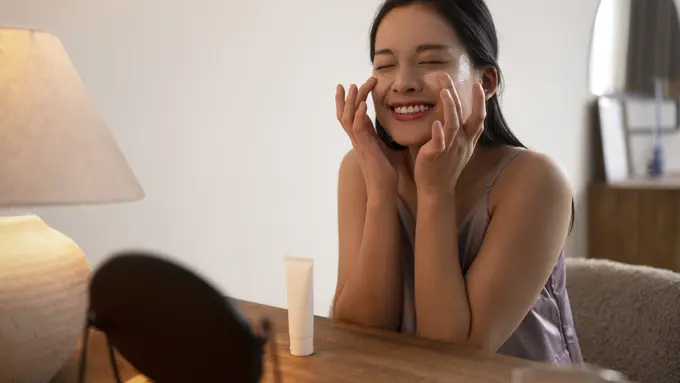 Seorang perempuan sedang mengenakan krim wajah sambil berbermin (Foto Dok: Freepik/freepik)