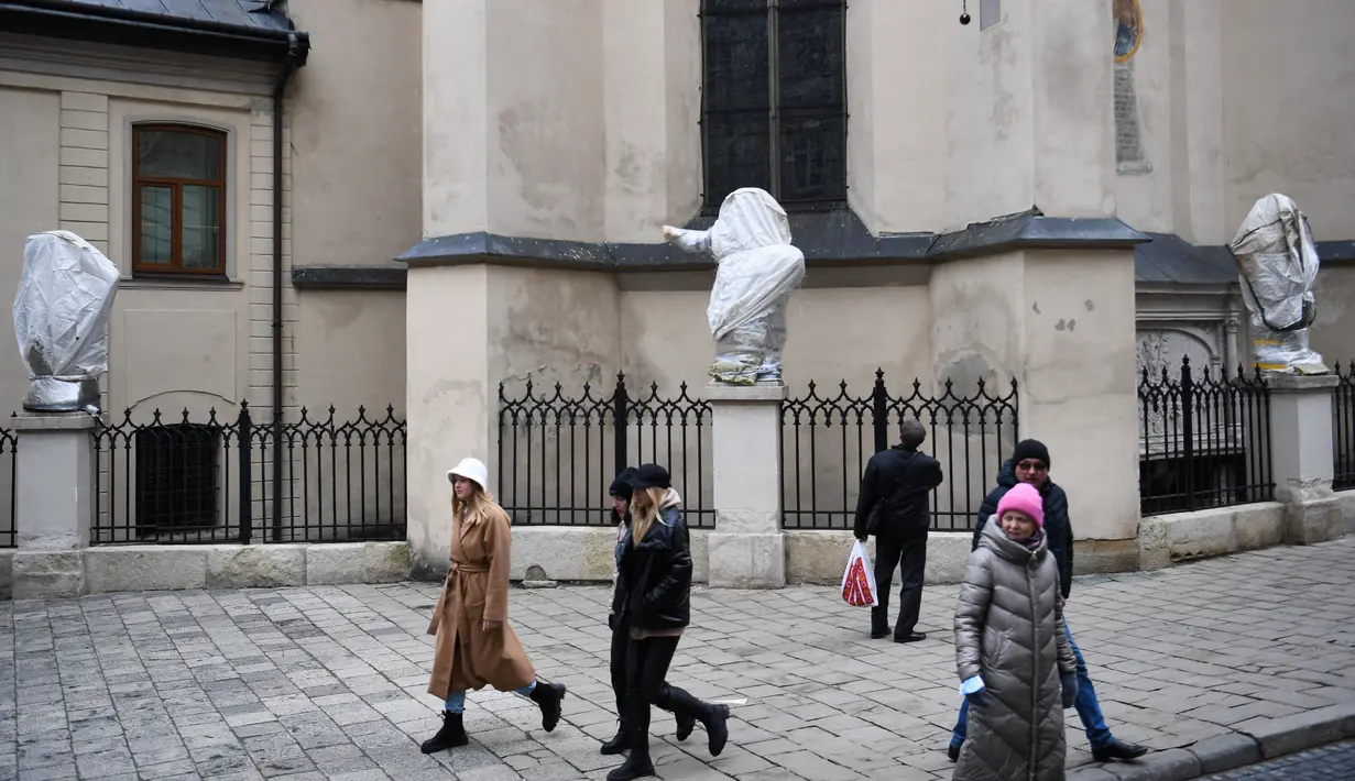 Pejalan kaki berjalan melewati patung-patung yang dibungkus dengan plastik di Katedral Latin di Lviv, 5 Maret 2022. Warga kota Lviv membungkus semua patung yang ada di kota tersebut untuk melindungi warisan sejarahnya dari kerusakan akibat serangan di tengah konflik Rusia-Ukraina. (Daniel LEAL/AFP)