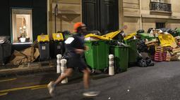 Seorang pria berolahraga melewati tong sampah yang tidak terkumpul di Paris, Prancis, Senin (13/3/2023). Demonstrasi besar-besaran serta pemogokan bergilir di Paris dan kota-kota lain untuk menolak peraturan pensiun itu telah berdampak pada kilang, transportasi umum dan pengumpulan sampah. (AP Photo/Michel Euler)