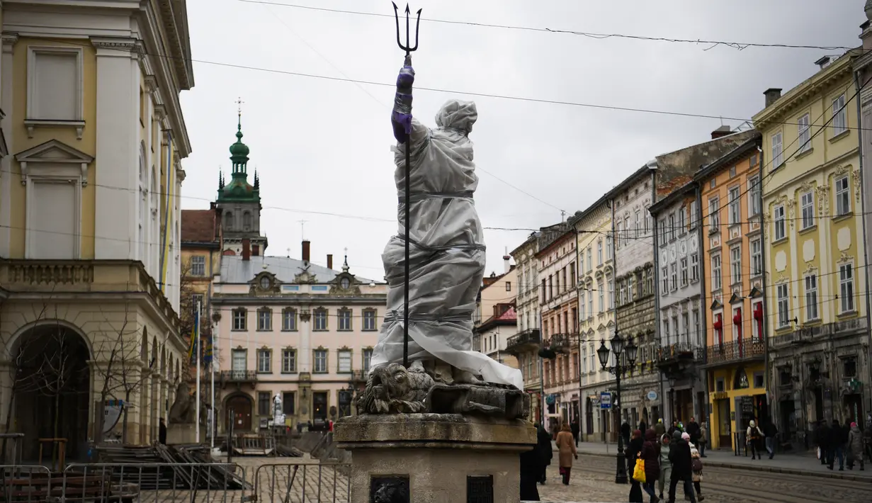 Pejalan kaki berjalan melewati patung yang dibungkus dengan plastik pelindung di sebelah Katedral Latin di Lviv, 5 Maret 2022. Warga kota Lviv membungkus semua patung untuk melindungi warisan sejarahnya dari kerusakan akibat serangan di tengah konflik Rusia-Ukraina. (Daniel LEAL/AFP)