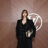 Lisa Blackpink hadir dengan gaya serba hitam mengenakan coat panjang dengan inner jumpsuit berkancing gold dipadukan kalung panjang tanpa liontin dan sepatu boots heels hitam. Ia membawa handbag warna coklat. [dok. Louis Vuitton]