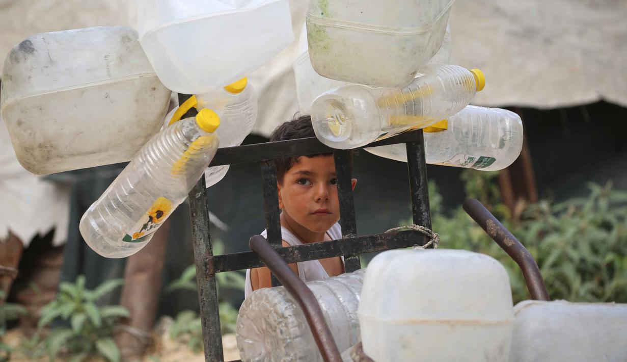 Seorang anak laki-laki Palestina berdiri di samping wadah plastik di atas troli, saat ia dan yang lainnya mengumpulkan air minum untuk keluarganya yang mengungsi dan tinggal di tempat penampungan setelah rumah-rumah mereka hancur akibat bombardir Israel, di kamp pengungsi Bureij, Jalur Gaza Tengah pada Senin 18 Mei 2026. (Eyad Baba/AFP)