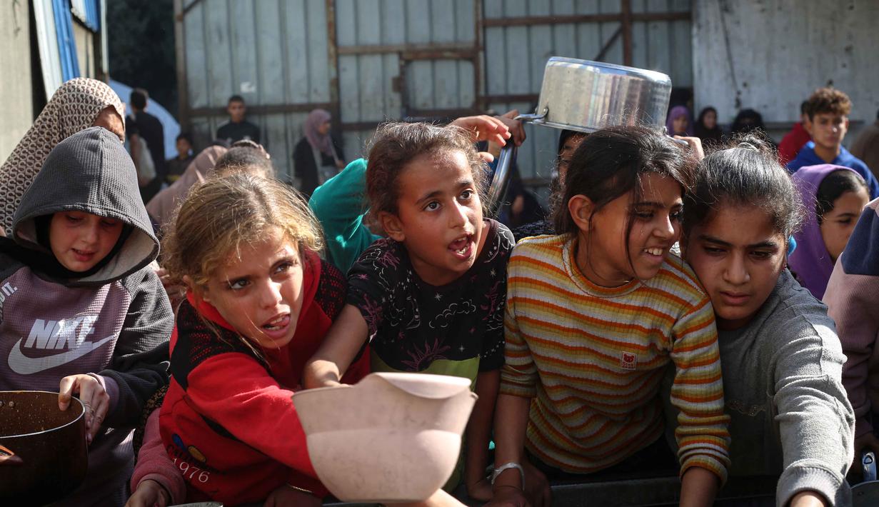 Terhambatnya peningkatan bantuan ke Gaza disebabkan beberapa faktor di antaranya, pembatasan visa, persetujuan impor, minimnya titik penyeberangan, serta berbagai hambatan teknis dan administratif lainnya. Tampak dalam foto, warga Palestina yang terlantar, anak-anak, dan dewasa, menunggu makanan di sebuah tempat penampungan yang telah dihuni keluarga-keluarga di Nuseirat, pusat Jalur Gaza, pada 23 November 2025. (EYAD BABA/AFP)