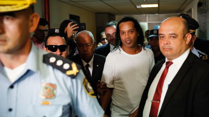 Mantan pemain timnas Brasil, Ronaldinho dikawal oleh polisi meninggalkan kantor pengadilan di Asuncion, ibu kota Paraguay, Jumat (6/3/2020). Ronaldinho bersama saudara laki-lakinya berurusan dengan pihak berwenang Paraguay karena menggunakan paspor palsu untuk memasuki negara itu. (AP/Jorge Saenz)