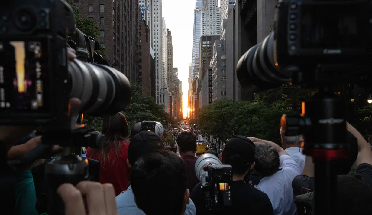 Orang-orang mengambil gambar saat matahari terbenam di Manhattan ketika fenomena "Manhattanhenge" di 42nd street, New York, Senin (11/7/2022). Fenomena Manhattanhenge hanyaterjadi di musim panas, tepatnya diantara bulan Mei hingga Juli. (Yuki IWAMURA / AFP)