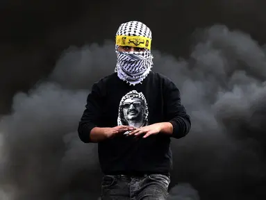 Seorang pemuda Palestina mengenakan kemeja bergambar mendiang pemimpin Yasser Arafat berjalan melewati ban yang terbakar saat konfrontasi dengan pasukan keamanan Israel menyusul demonstrasi menentang perampasan tanah oleh Israel di Desa Kfar Qaddum, Tepi Barat, 4 Maret 2022. (JAAFAR ASHTIYEH/AFP)
