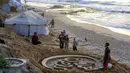 Meski bersifat sementara karena sering terhapus oleh pasang air laut di malam hari, namun Yazeed Abu Jarad bersama timnya secara rutin kembali setiap pagi untuk membuat karya baru sebagai simbol harapan. Tampak dalam foto, warga membantu seniman Palestina Yazeed Abu Jarad, saat ia membuat patung pasir dengan pesan ucapan "Selamat Datang, Ramadan" di sepanjang pantai di Khan Younis, selatan Gaza pada 17 Februari 2026. (Bashar Taleb/AFP)