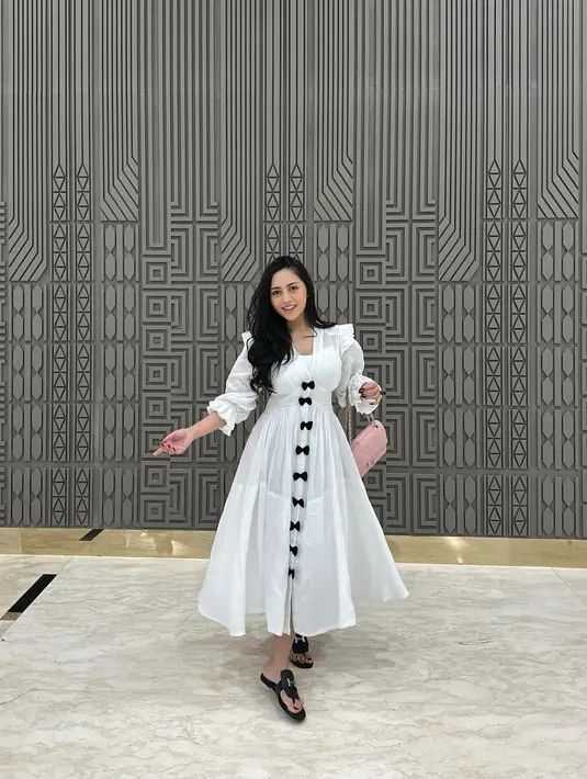 Penampilan manis lainnya Rachel Vennya pakai dress putih. Dress ini memiliki detail lengan Panjang dengan kancing berbentuk pita-pita kecil hitam hingga ujung bawah. Rachel memadukan penampilannya dengan tas pink dan sandal hitam. [Foto: Instagram/rachelvennya]
