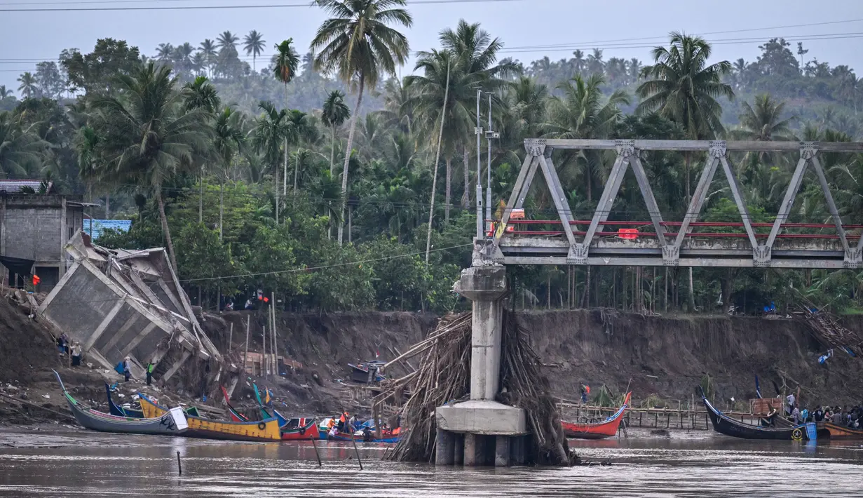 Sisa rangka jembatan terlihat menggantung di atas aliran sungai Peusangan yang masih deras. Tampak dalam foto, perahu-perahu yang digunakan untuk mengangkut penumpang menyeberangi Sungai Peusangan terlihat melewati jembatan yang runtuh di Kuta Blang, Kabupaten Bireuen, Provinsi Aceh, Selasa 9 Desember 2025. (CHAIDEER MAHYUDDIN/AFP)