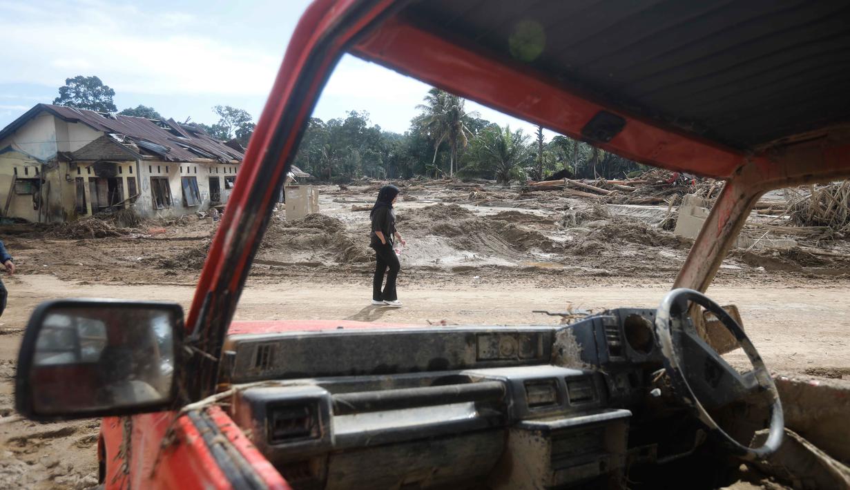 Kerusakan mencakup rumah penduduk, jalan, irigasi, persawahan, sekolah, jaringan air bersih, serta fasilitas umum lainnya. Tampak dalam foto, seorang perempuan terlihat di antara reruntuhan mobil saat berjalan di permukiman yang terdampak banjir bandang di Batang Toru, Sumatera Utara, Senin 1 Desember 2025. (AP Photo/Binsar Bakkara)