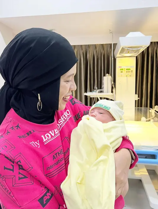 Geni Faruk biasa tampil colorful, termasuk ketika momong cucu. Potret gemas Geni Faruk dengan atasan berwarna pink dengan hijab hitam menggendong anak kedua Aurel dan Atta. [Foto: Instagram/genifaruk]