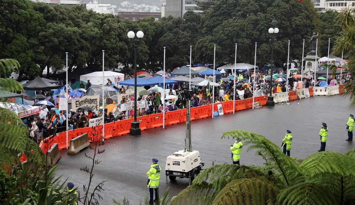 Para pengunjuk rasa (kiri) memadati halaman Parlemen ketika polisi (kanan) mengawasi pada hari kelima demonstrasi menentang pembatasan COVID-19 di Wellington, Selandia Baru, 12 Februari 2022. Aksi ini terinspirasi oleh demonstrasi serupa di Kanada. (MARTY MELVILLE/AFP)