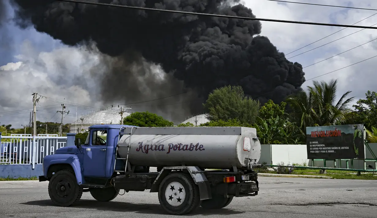 Asap hitam dari kebakaran besar di depot bahan bakar terlihat saat sebuah truk air melewati jalan di Matanzas, Kuba (7/8/2022). Kuba meminta bantuan Sabtu (7/7) untuk mengatasi kebakaran besar yang menyebabkan 77 orang terluka dan 17 petugas pemadam kebakaran hilang. Sekitar 800 orang telah dievakuasi dari daerah tersebut. (AFP/Yamil Lage)