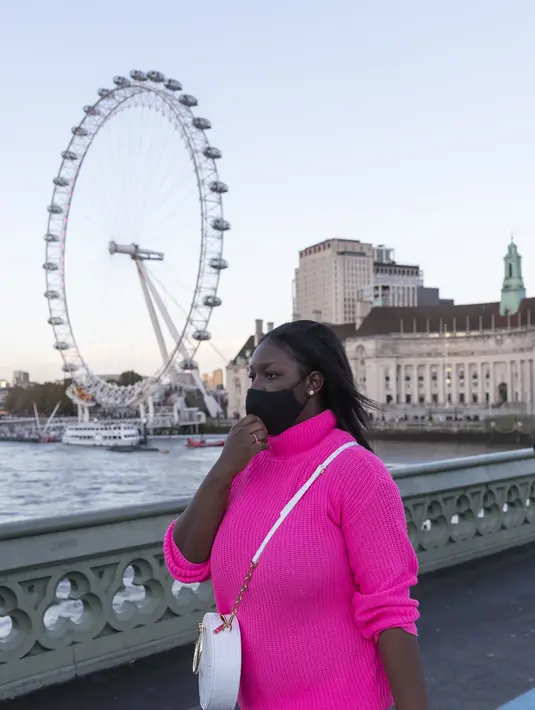 Seorang wanita yang mengenakan masker berjalan di Jembatan Westminster dengan latar pemandangan London Eye di London, Inggris (31/10/2020). Kasus baru COVID-19 di Inggris mencapai 21.915, menambah total kasus coronavirus di negara itu menjadi 1.011.660. (Xinhua/Han Yan)