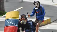 Pengunjuk rasa berada di balik tameng saat mereka bentrok dengan polisi antihuru-hara menuntut Presiden Venezuela, Nicolas Maduro, di Caracas (10/7). Salah satu demonstran mengenakan pelindung wajah karakter Mickey Mouse. (AFP Photo/Juan Barreto)