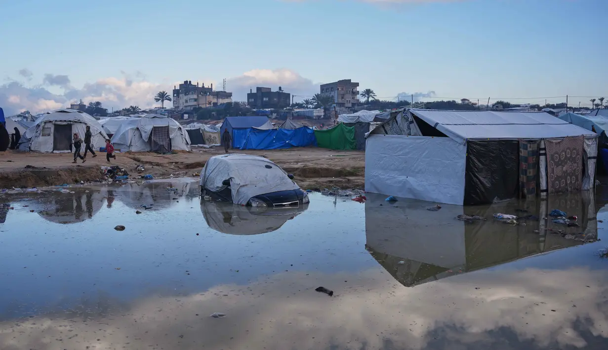 Banjir juga memicu risiko penyakit menular akibat sistem drainase yang hancur akibat serangan Israel sejak Oktober 2023 lalu. Tampak dalam foto, anak-anak Palestina berlari melewati sebuah mobil yang sebagian terendam di daerah yang tergenang banjir setelah hujan lebat di sebuah kamp pengungsian sementara, Zawaida, Jalur Gaza tengah, Rabu 17 Desember 2025. (AP Photo/Abdel Kareem Hana)