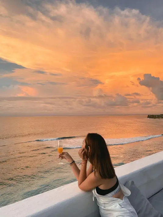 Menikmati minuman di pinggir pantai saat matahari terbenam, Pamela Bowie terlihat mengenakan sport bra yang ditumpuknya dengan jumpsuit tanpa lengan berwarna putih. Foto: Instagram.