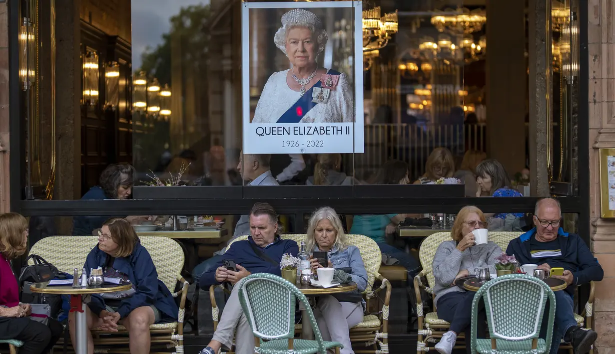 Orang-orang duduk di bar teras di sebelah potret Ratu Elizabeth II di pusat kota London pada 13 September 2022. Hotel, restoran, dan toko-toko penuh sesak saat penggemar kerajaan berdatangan ke jantung kota London untuk melihat jalan-jalan yang dipagari bendera, prosesi yang dipenuhi kemegahan, dan berani mengantre sejauh bermil-mil untuk kesempatan sekali seumur hidup mengucapkan selamat tinggal kepada Ratu Elizabeth II. (AP Photo/Emilio Morenatti, File)