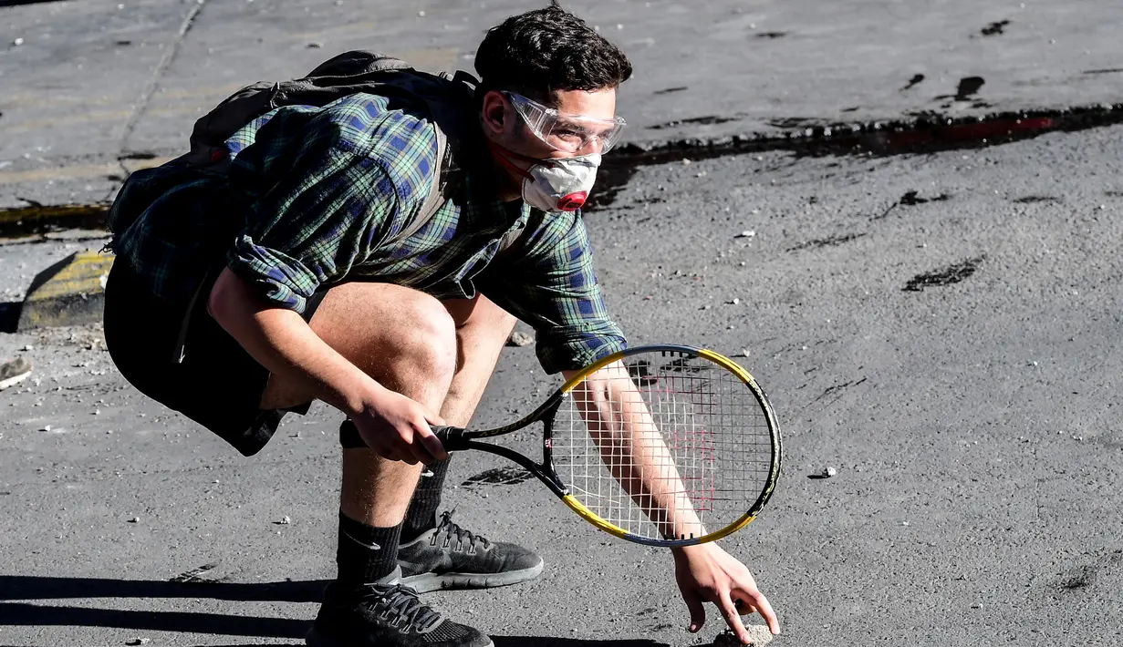 Seorang demonstran membawa raket tenis mengambil batu saat bentrok dengan polisi anti huru hara selama memprotes kebijakan ekonomi pemerintah di Santiago (6/11/2019). para pemrotes meminta para demonstran untuk memperluas aksi unjuk rasa. (AFP Photo/Martin Bernetti)
